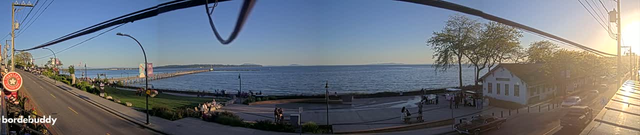 A panoramic view of a waterfront town on a sunny day, showing a long pier extending into calm water, a road with some pedestrians, and buildings along the shore, with trees and a sunset in the background.