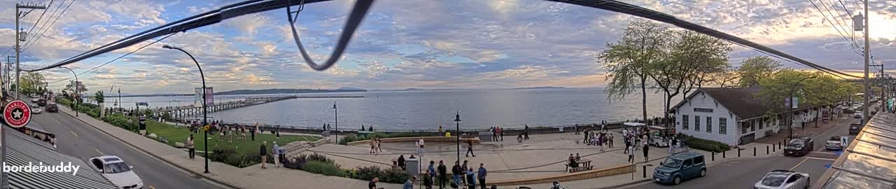 A waterfront scene under a partly cloudy sky shows a town square with people gathered near a pier extending into a calm body of water, with cars parked along a street and trees lining the shoreline.