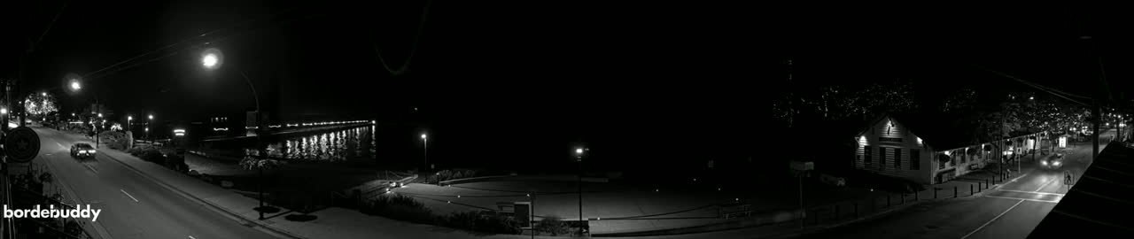 A nighttime, black-and-white view of a street scene with cars, buildings, and a waterfront pier, all illuminated by streetlights.