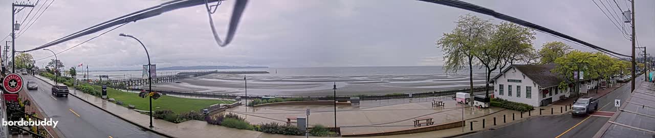 A wet, overcast day reveals a coastal town scene with a long pier extending into a calm ocean, a paved plaza with picnic tables, and a street lined with trees and buildings.