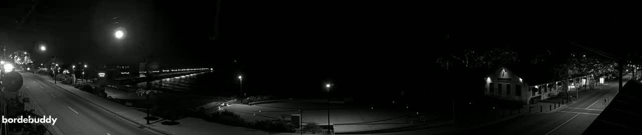 A nighttime, black-and-white panoramic view of a city street, a waterfront with lights lining a pier, and buildings, all under a clear, dark sky.