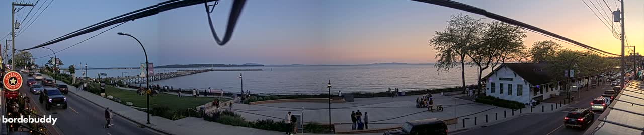 A panoramic view of a waterfront town at sunset, showing a calm body of water, a pier, a street with cars, and people gathered in a plaza near a white building.
