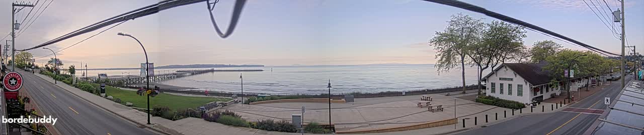 A seaside town at dawn, with a calm ocean, a long pier, and a paved plaza near a white building next to a quiet street.