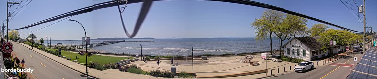 A sunny seaside town scene shows a calm ocean with a long pier, a grassy park, a paved plaza with picnic tables, a street with cars, and buildings under a clear blue sky.