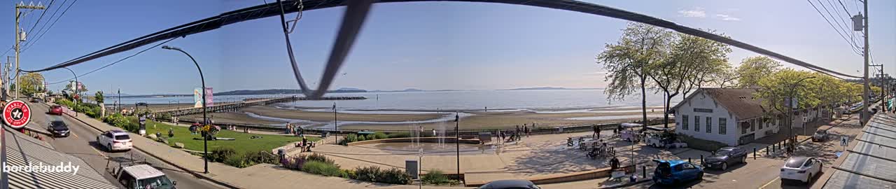 A sunny day at a seaside town, featuring a beach with a pier, a water fountain in a plaza, and parked cars along a street lined with trees and buildings.