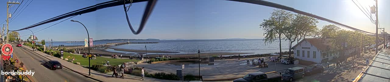 A sunny seaside town scene shows a long pier extending into a calm bay, with a street, buildings, and people visible along the shore.