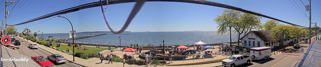 A sunny seaside town scene features a long pier extending into the water, a grassy area with people and market stalls, and parked cars along a street lined with trees and buildings.