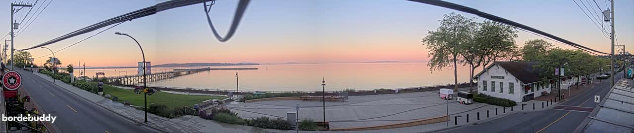 A panoramic view at sunrise shows a calm body of water with a pier, a waterfront town with a street and buildings, and a grassy area.