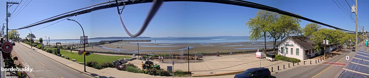 A sunny day overlooks a waterfront town with a pier, sandy beach, and a small plaza featuring a water fountain, all under a clear blue sky.