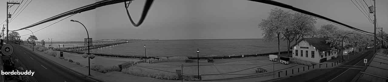 A grayscale panoramic view shows a calm lake with a long pier extending into it, a road alongside the water's edge with a building and trees, under a clear, possibly twilight sky.