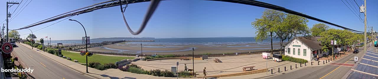 A sunny seaside town scene shows a wide sandy beach at low tide, a long pier, a street lined with trees and buildings, and people walking along the shore.