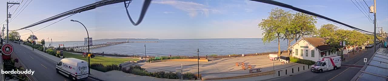 A waterfront scene on a sunny day shows a long pier extending into a calm body of water, a street with a few vehicles, and a small building near a park-like area with picnic tables.
