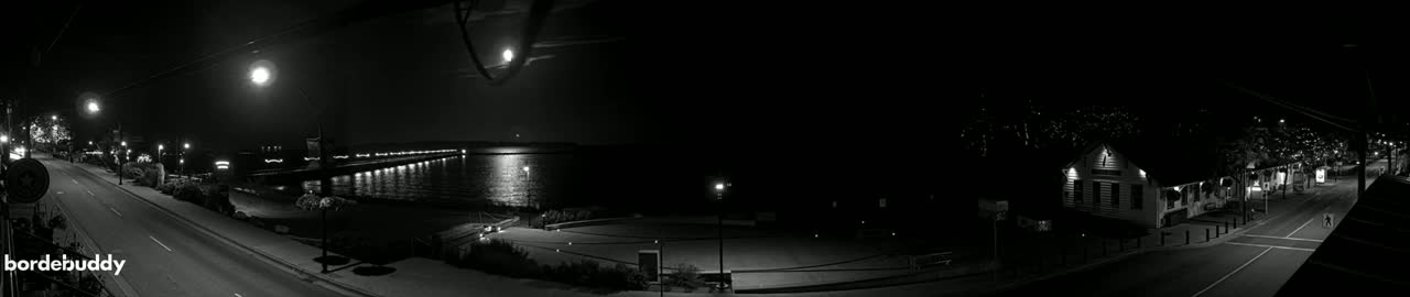 A nighttime panoramic view of a town's waterfront, showing a street leading to a pier illuminated by streetlights, with a calm body of water reflecting the lights under a mostly clear sky.
