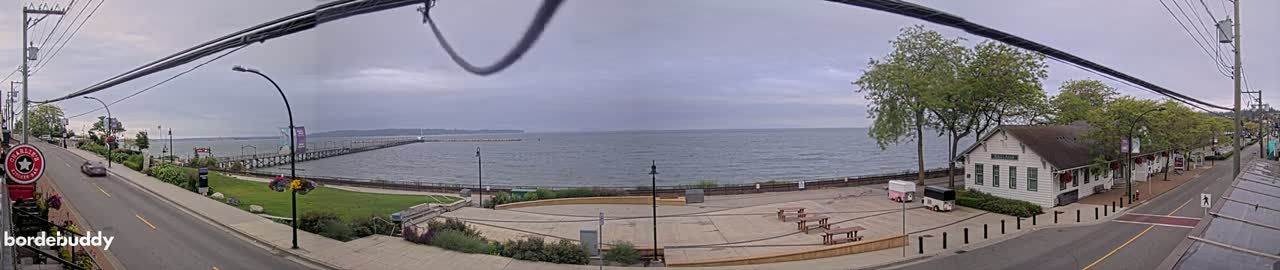 A waterfront scene on an overcast day shows a paved plaza with picnic tables near a white building and a road, all overlooking a calm body of water and a pier extending into the distance.
