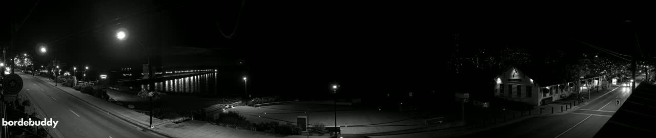 A nighttime, black and white panorama of a street scene with buildings, streetlights, and a waterfront pier illuminated.