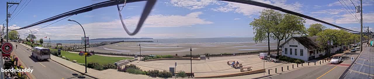 A sunny day reveals a seaside town with a sandy beach, a pier, and a white building near the water, a bus driving on the street, and cars parked along the side of the road.