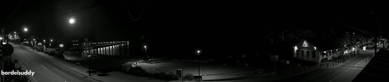 A nighttime, black-and-white panoramic view shows a quiet street and waterfront, with buildings and streetlights illuminated against a dark sky.
