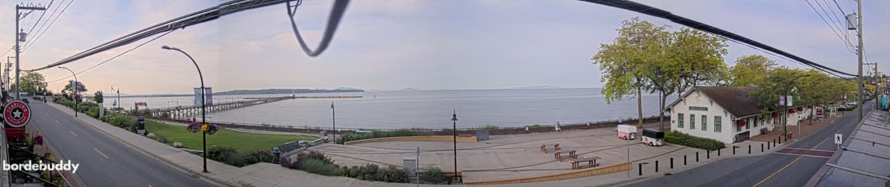 A seaside town on a partly cloudy day features a long pier extending into calm water, a paved waterfront area with picnic tables, a white building, and a street lined with trees.