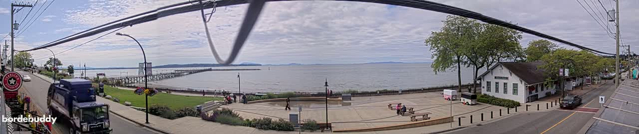 A seaside town scene on an overcast day features a calm bay, a long pier, a grassy park with people, a white building, and a street with a garbage truck.