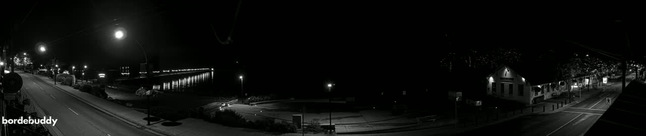 A nighttime, black and white panoramic view of a street scene shows a road leading to a pier, with buildings and trees lining the street under dim streetlights.
