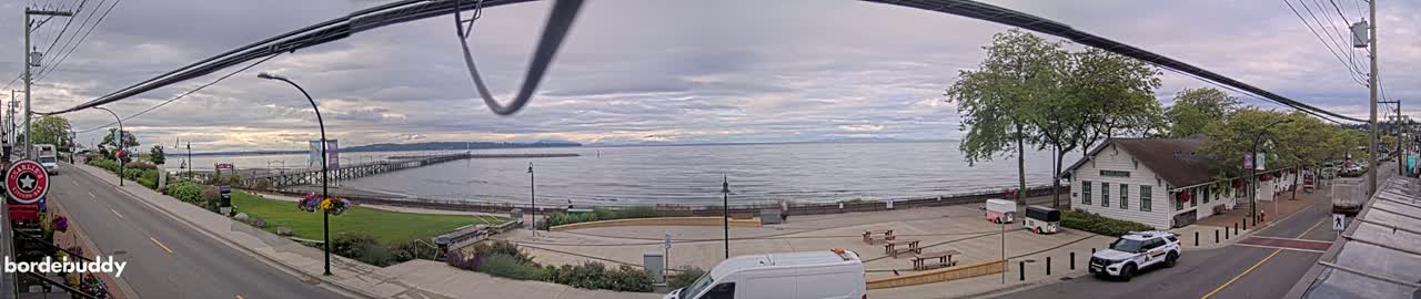 A waterfront scene on an overcast day shows a pier extending into a calm bay, a street with parked cars, and buildings along the shore, including a white building that resembles a small train station.