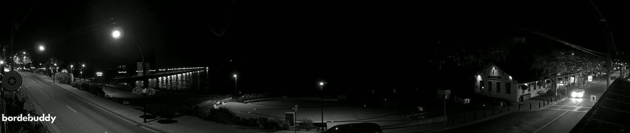 A nighttime, black and white view of a street scene with a lit-up pier in the distance, a building to the right, and cars driving on a road.