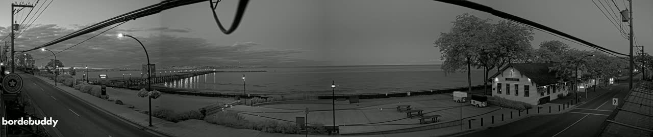 A black and white panoramic view shows a calm body of water at twilight, a long pier extending into it, a road with streetlights, and a small building near trees.
