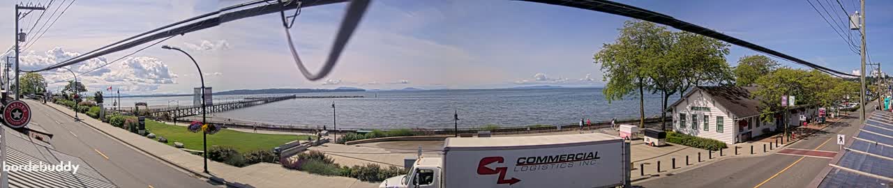 A seaside town scene on a partly sunny day shows a long pier extending into a calm body of water, with a park, buildings, and a road visible in the foreground.