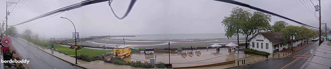 A wet, overcast seaside town scene shows a street, a pier, a beach, and a building with tents, all under a gray sky.