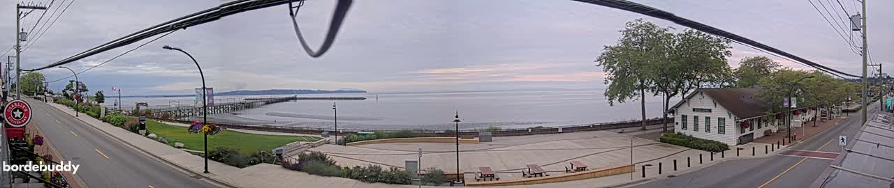 A seaside town scene on an overcast day features a long pier extending into calm water, a paved waterfront area with picnic tables, a white building, and a street with overhead power lines.