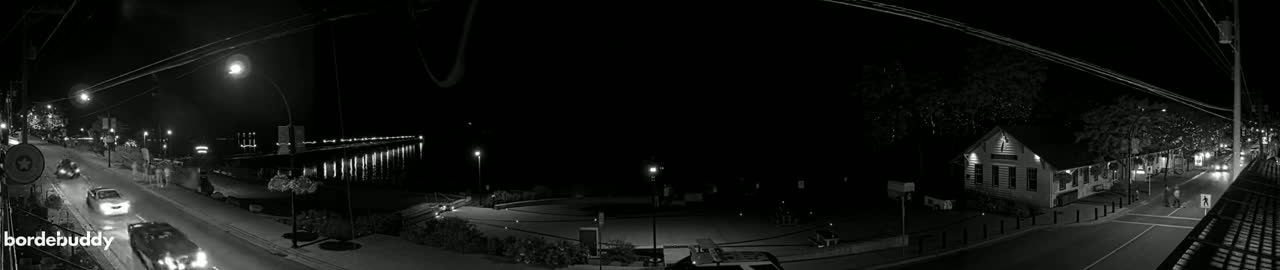 A nighttime, black and white view of a town's waterfront, featuring a pier with lights, a roadway with cars, and buildings under a clear, dark sky.