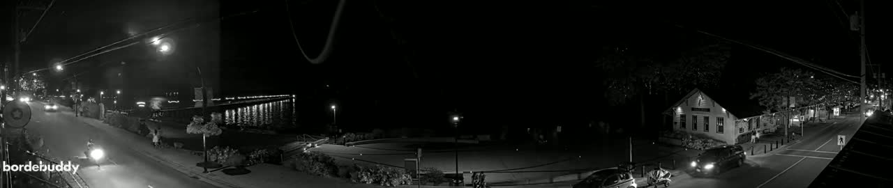 A nighttime, black-and-white view of a town's waterfront at night, showing a street with cars, a pier with lights reflecting on the water, and buildings along the street.