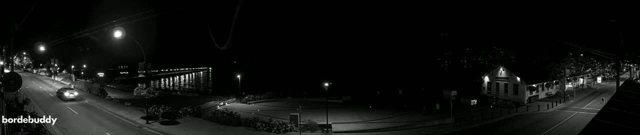 A nighttime, black-and-white view of a street scene with cars driving on a road that passes by a waterfront pier and a building with lights on.