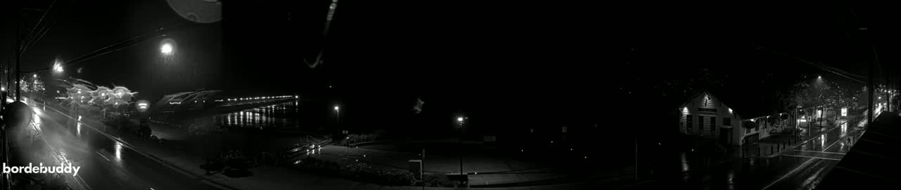 A nighttime, black-and-white view of a street scene with cars driving on a road that passes by a waterfront pier and a building with lights on.