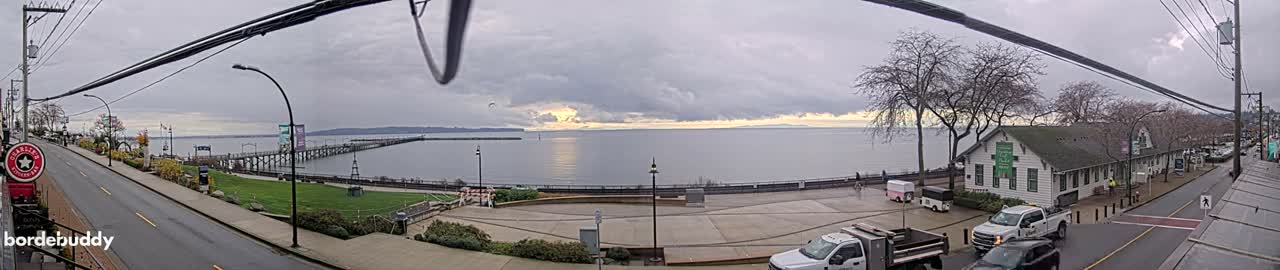 A nighttime, black-and-white view of a street scene with cars driving on a road that passes by a waterfront pier and a building with lights on.