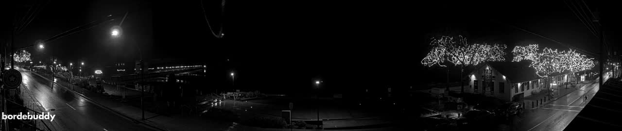 A panoramic black and white image depicts a dark, wet street at night, illuminated by streetlights, buildings, and trees adorned with bright decorative lights, under rainy conditions.