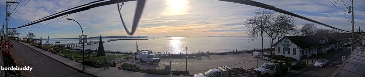 A panoramic view captures a sunny coastal town during golden hour, featuring a long pier extending into calm water, a prominent decorated Christmas tree by the shore, and a street lined with buildings and vehicles under a partly cloudy sky.