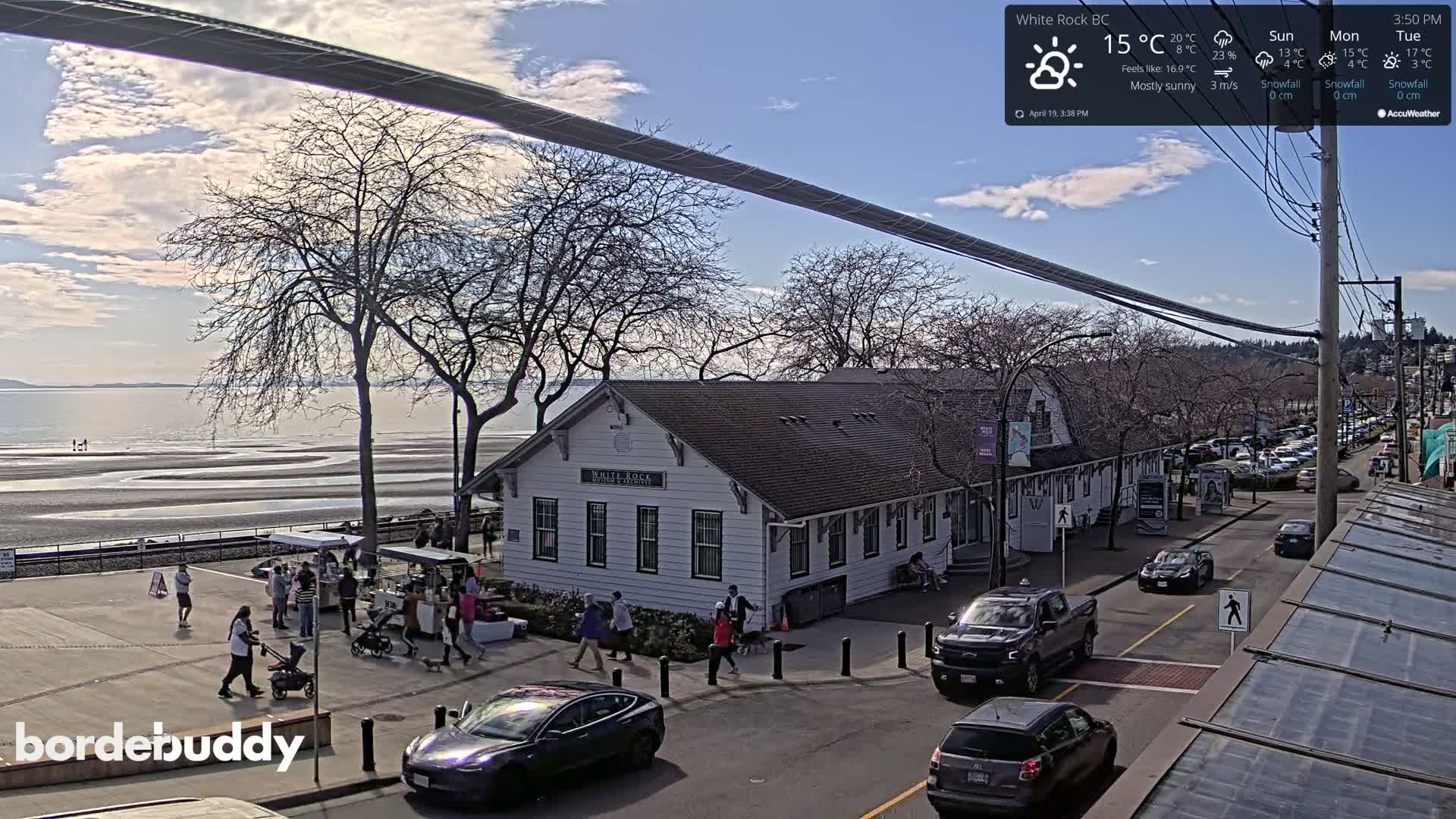 A mostly sunny seaside town scene shows a white building, pedestrians, cars, and a long cable strung overhead, with a calm ocean and beach in the background.