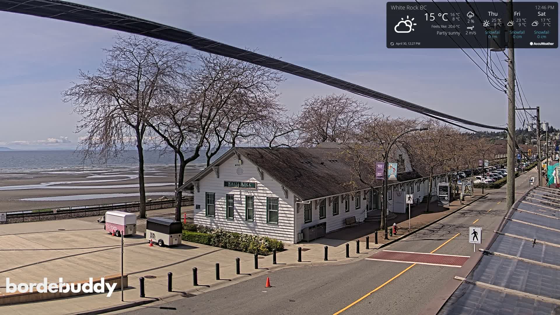 A partly sunny seaside scene shows a white building near a street with parked cars, a sandy beach, and calm ocean waters in the background.