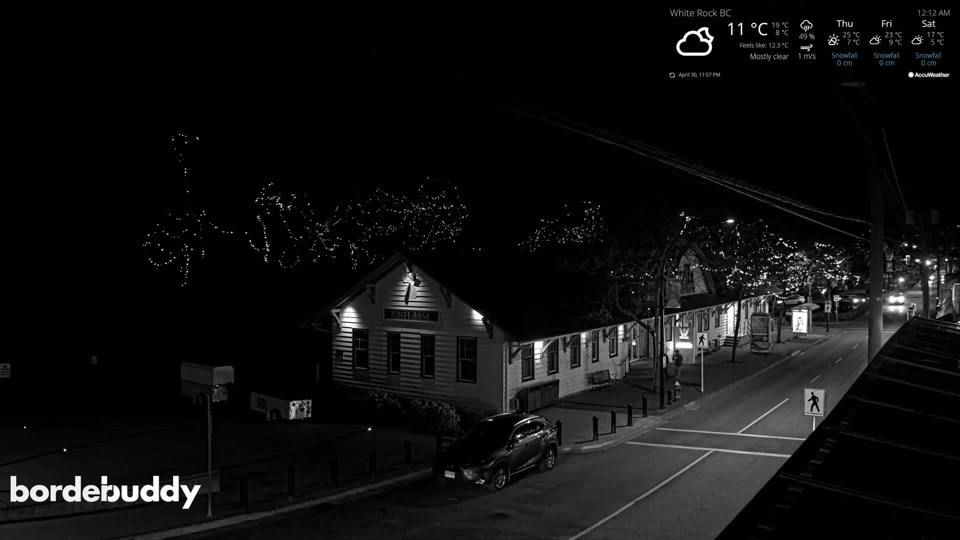 A nighttime, mostly clear street scene shows a two-story building with string lights overhead and a car parked in front of it.