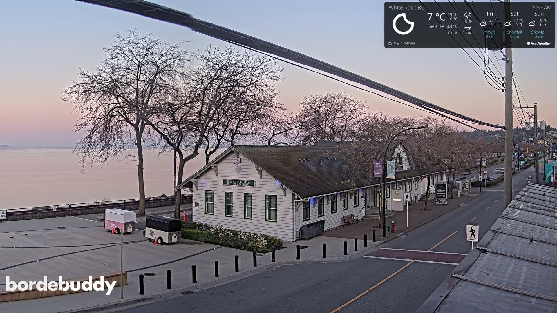 A white building sits beside a calm body of water under a pale pink and purple sunrise sky.