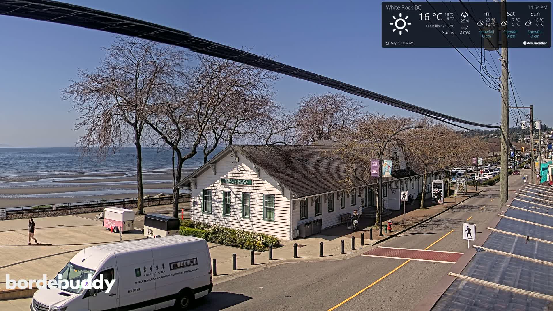 A sunny seaside town scene shows a white building near a calm ocean, a street with parked cars, and bare trees.