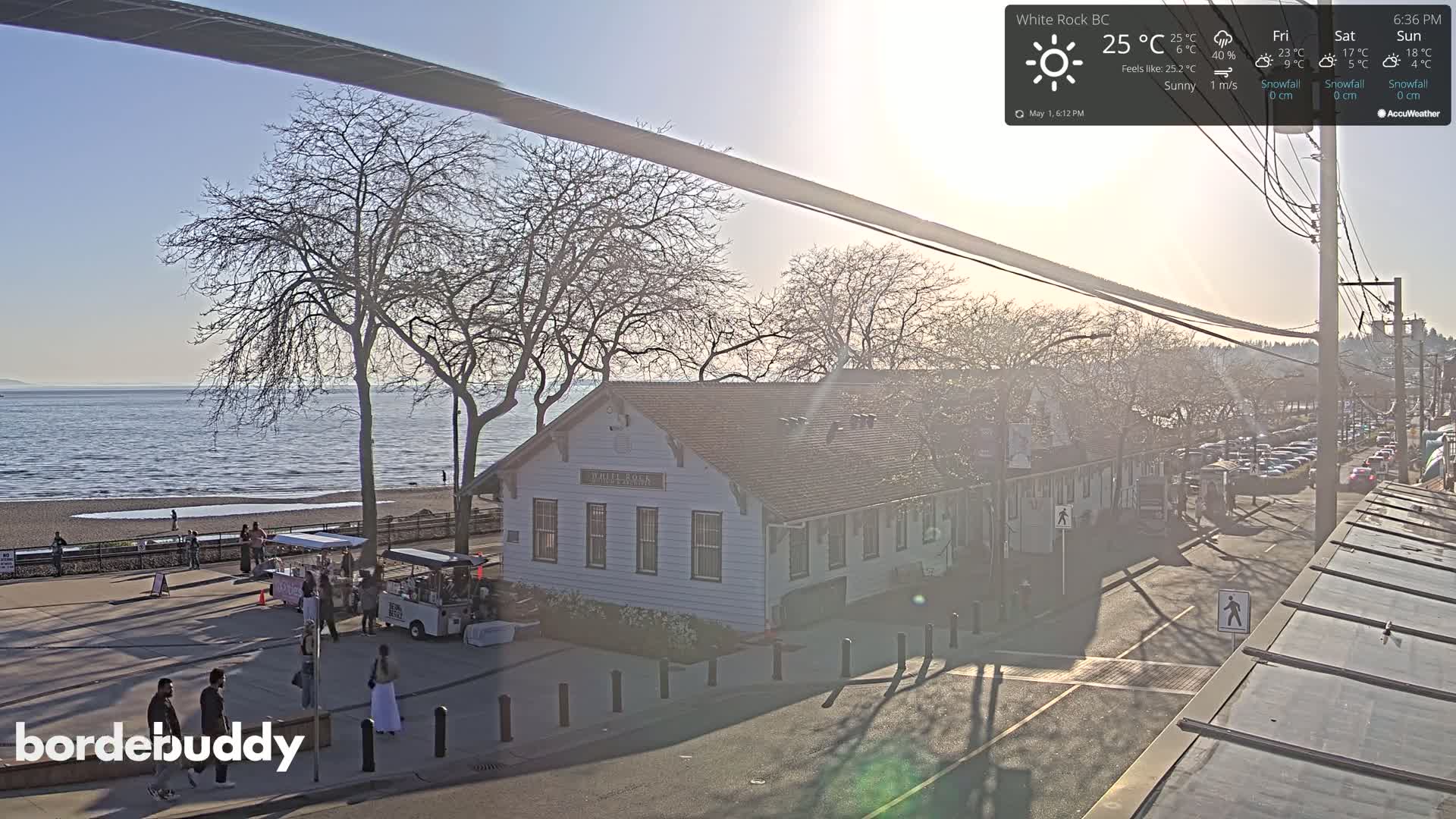 A sunny waterfront scene shows a building near a beach with people and food carts, a calm sea, and bare trees under a mostly clear sky.