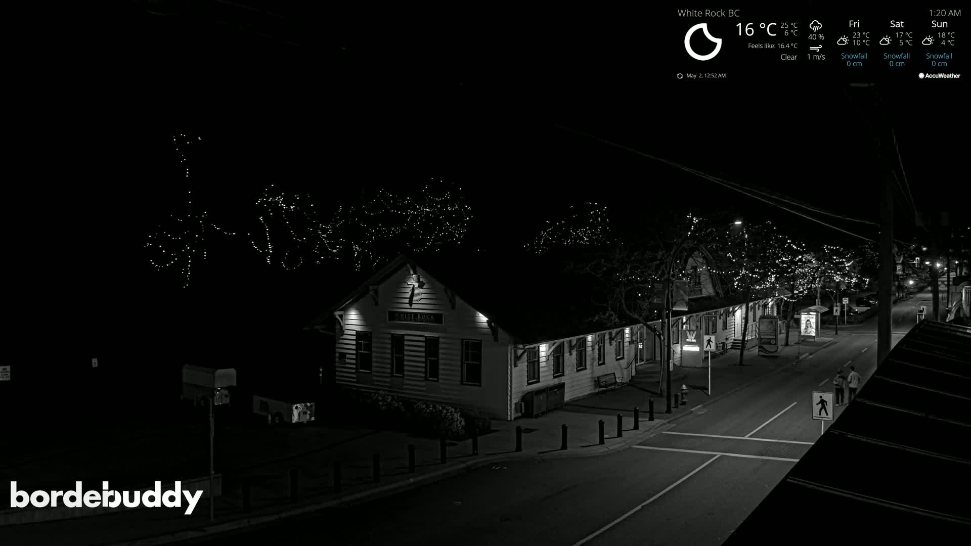 A nighttime, clear street scene shows a building with string lights on the trees, and a few pedestrians walking.