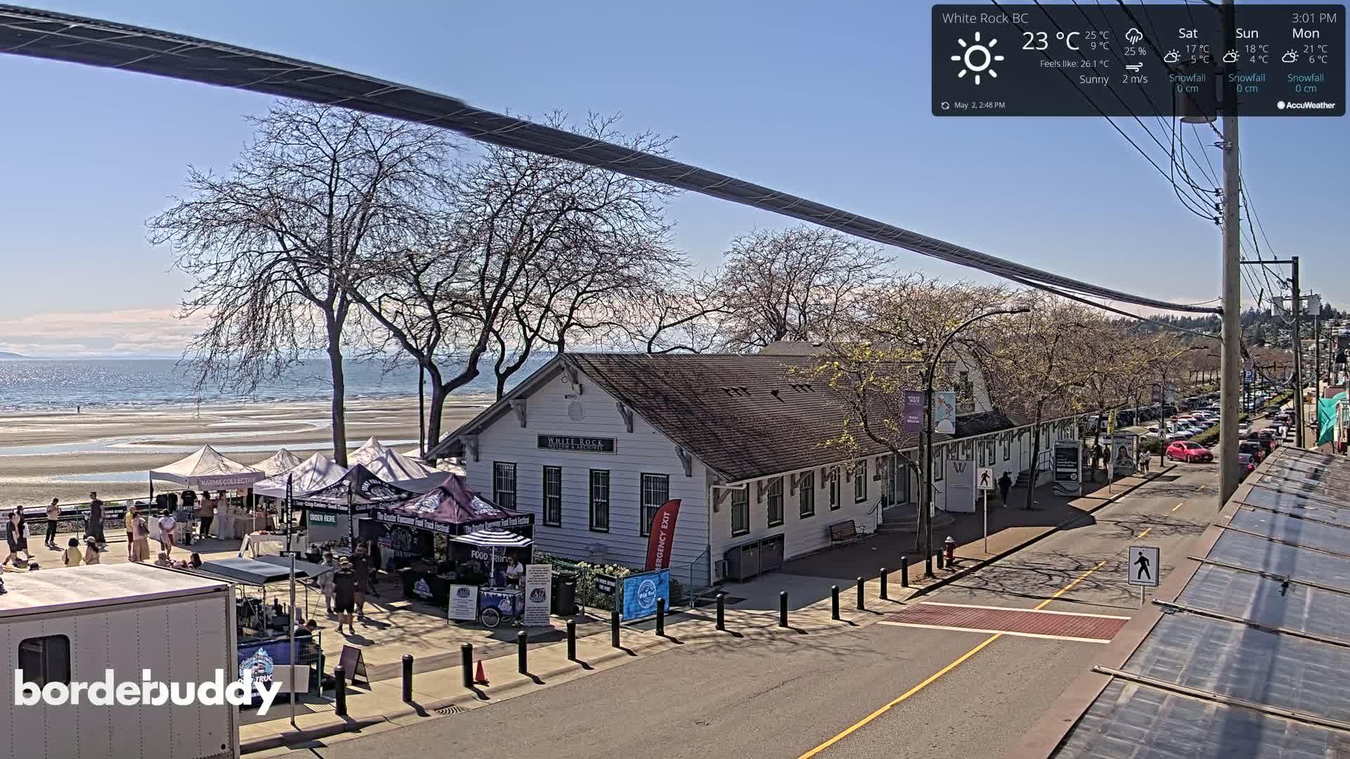 A sunny seaside town features a market with numerous tents near a white building, a calm ocean in the background, and a street with parked cars.