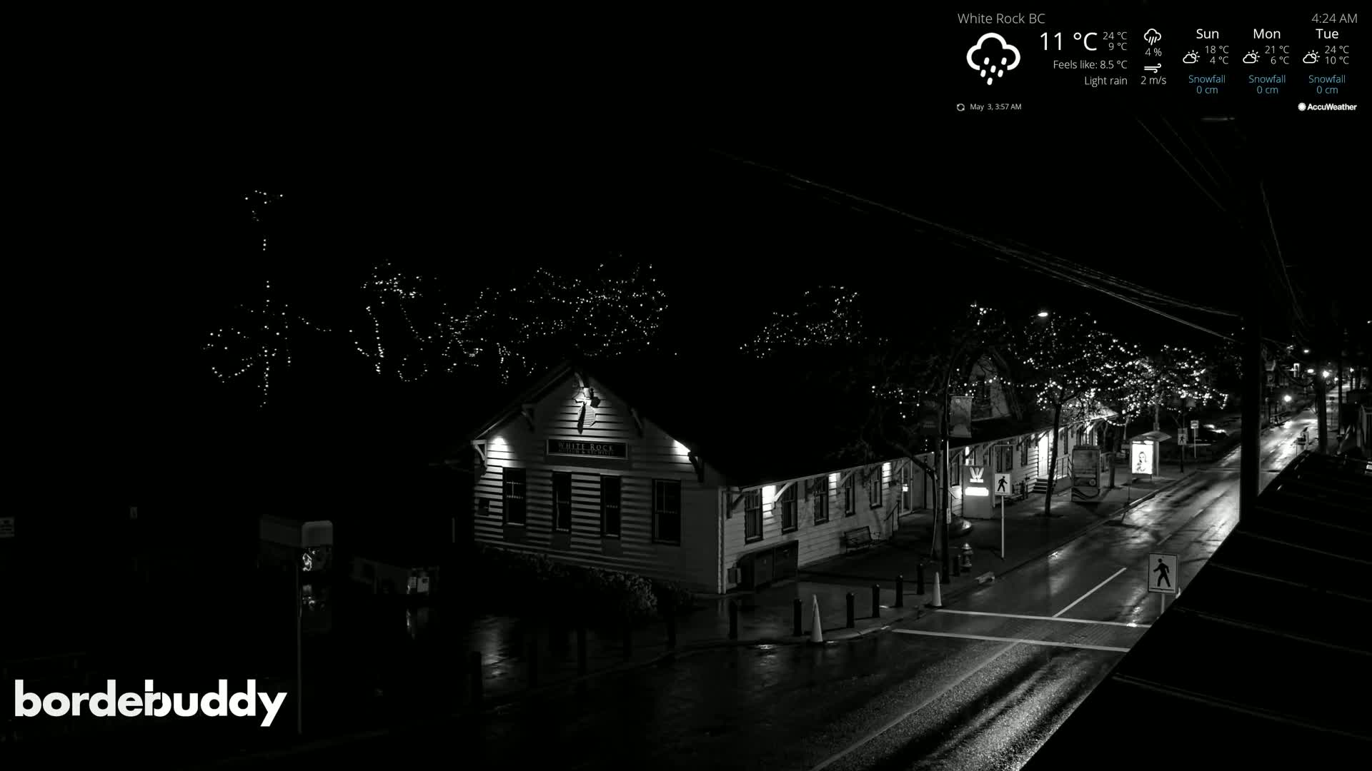 A rain-slicked street at night, lined with buildings decorated with string lights, is viewed from above.