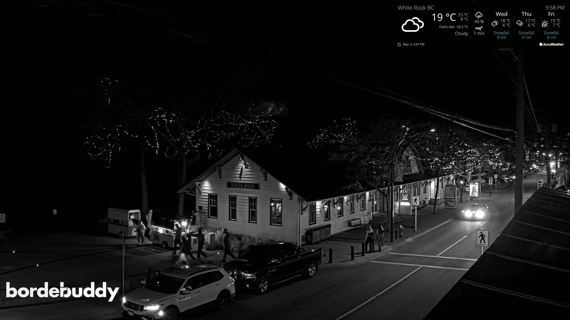 A nighttime black and white view of a street scene shows a building with string lights on nearby trees, several vehicles, and pedestrians walking along a roadway under a cloudy sky.
