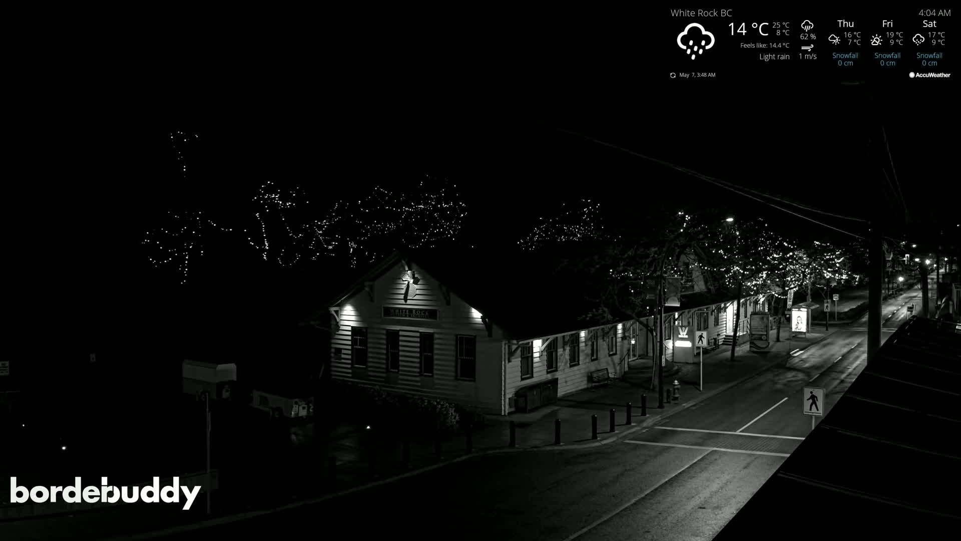 A nighttime, lightly rain-slicked street scene shows a building with lights on and string lights in the trees above.
