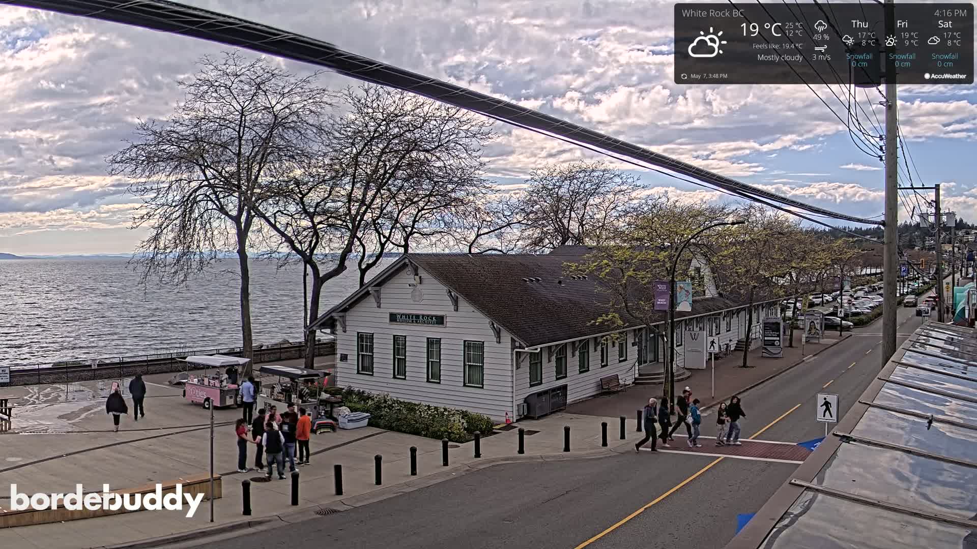 A mostly cloudy day overlooks a waterfront town with a white building, pedestrians, and parked cars.