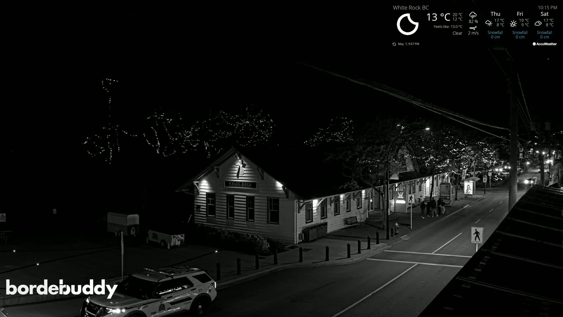 A nighttime, clear black and white view of a street scene shows a building with string lights on nearby trees, a police vehicle, and a few pedestrians.